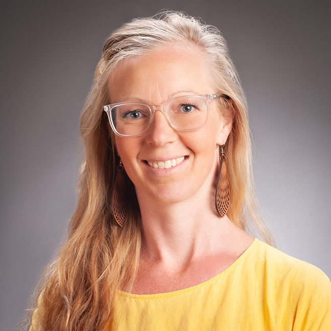 An image of Dr Eva Neely who has long blond hair and is wearing a yellow shirt and glasses.