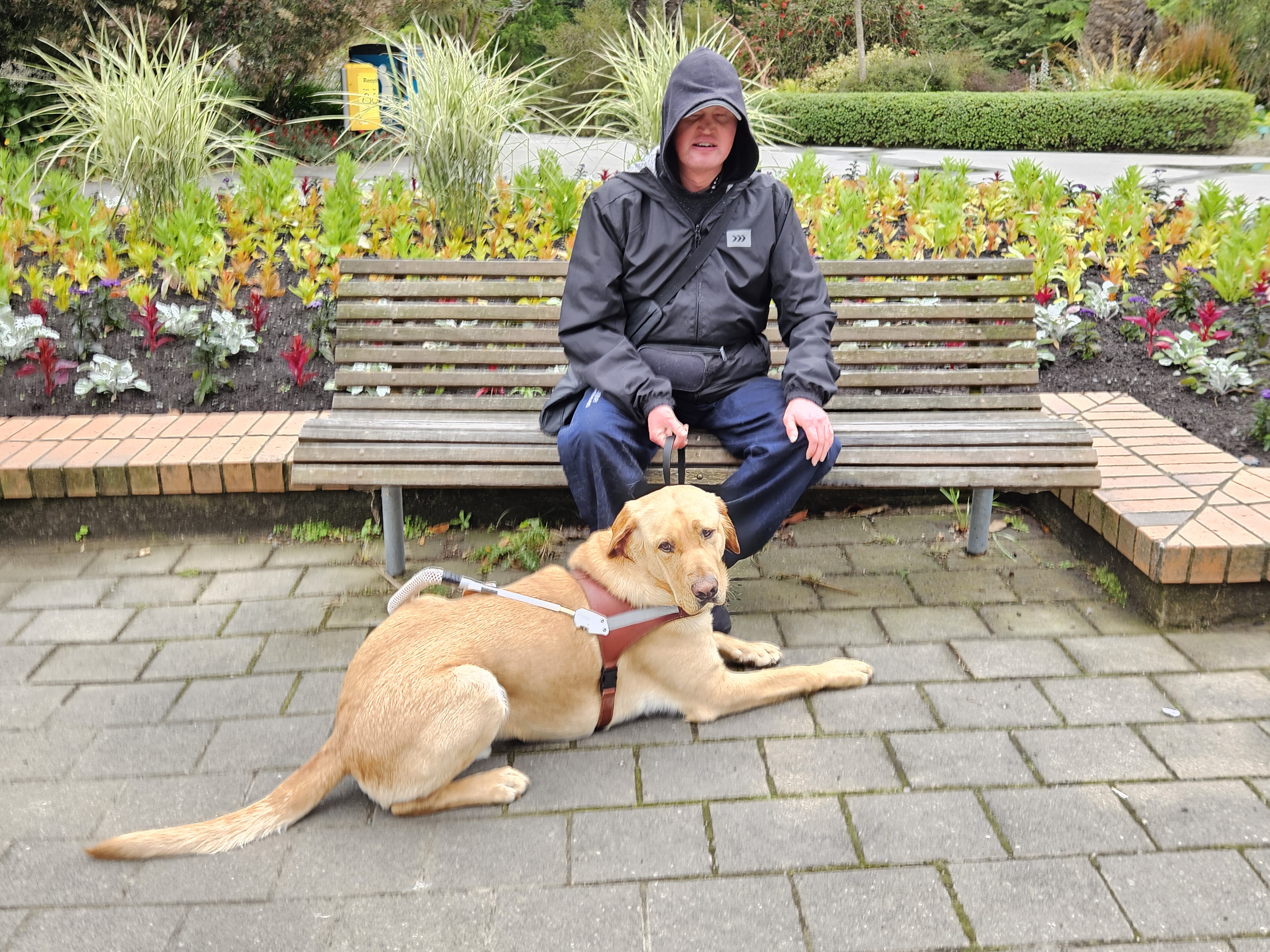 Middle aged man (Carl Lintott) wearing rain gear sits on a bench in a park with a yellow guide dog sitting at his feet. Background shows a garden with many green and red plants growing and a couple of rubbish bins in the background.